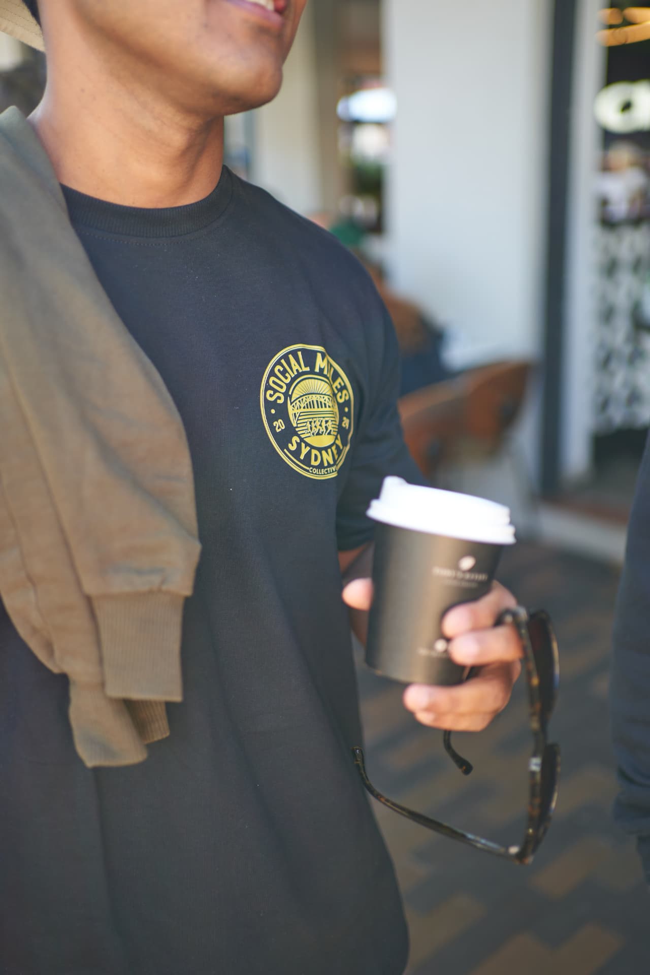 A Social Miles Sydney member in a club tee holding a takeaway coffee after the Sunday loop.